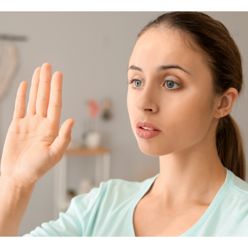 A woman with brown hair in a ponytail raises her hand, palm facing forward, against a blurred indoor background. She appears focused and attentive.