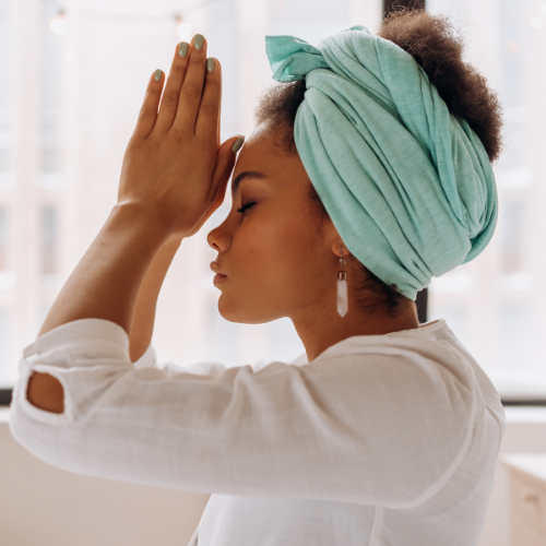 Person in white shirt and turquoise head wrap stands in profile with hands together, fingertips touching forehead, in a calm indoor setting.
