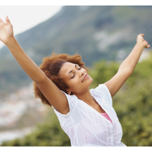 Person outdoors with arms raised, eyes closed, smiling, wearing a white shirt. Blurred greenery and hills in the background.