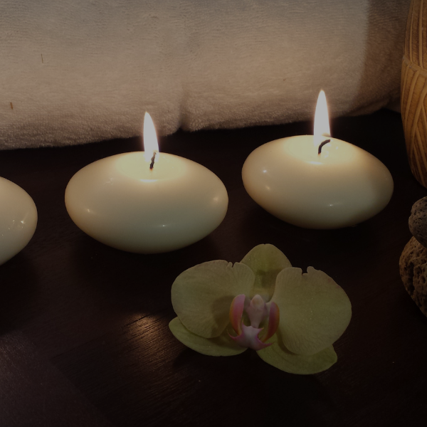 The Impact of Reiki on Depression Three lit, round candles on a dark surface cast a serene glow, complemented by a light green orchid in front. A towel rests in the background, creating a tranquil setting perfect for reiki practice and restoring energy balance.