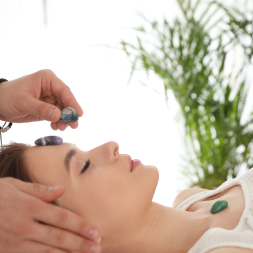 Person lying down with eyes closed, while another places crystals on their forehead and chest. A plant is visible in the background.