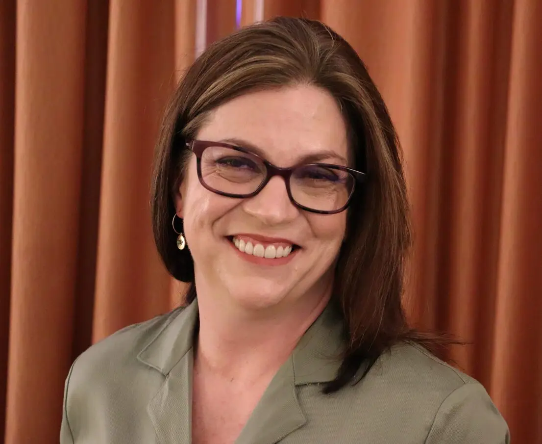 A person with brown hair and glasses, exuding a warm smile, dons a green blazer as they stand before brown curtains. Their presence radiates the calm energy of a reiki practitioner, inviting an aura of healing into the space.