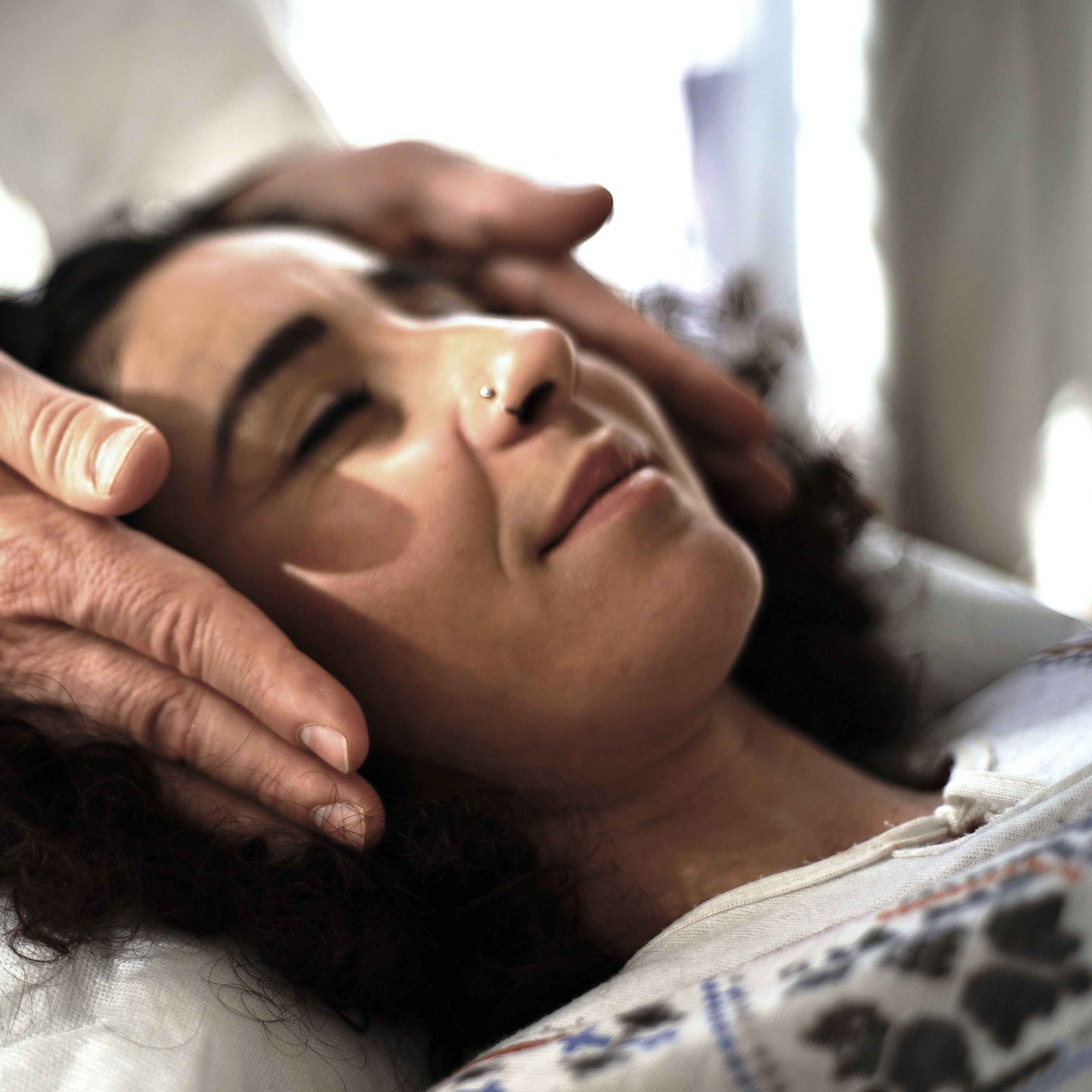 A person with curly hair is lying down, eyes closed, while another person's hands gently touch their head in a soothing reiki session.