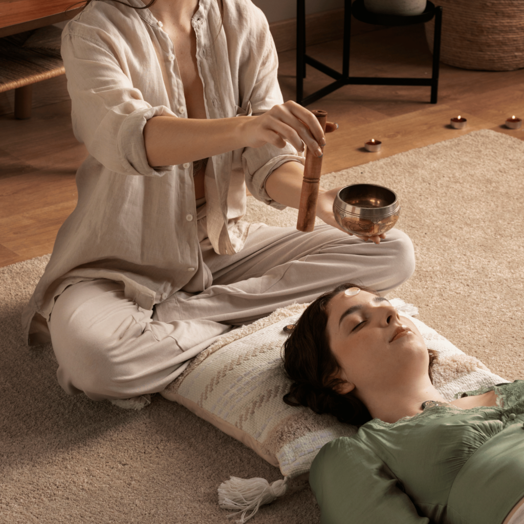 A person is lying down with eyes closed while another gently holds a singing bowl above them, creating a peaceful setting that complements the benefits of Reiki Therapy for chronic pain management.