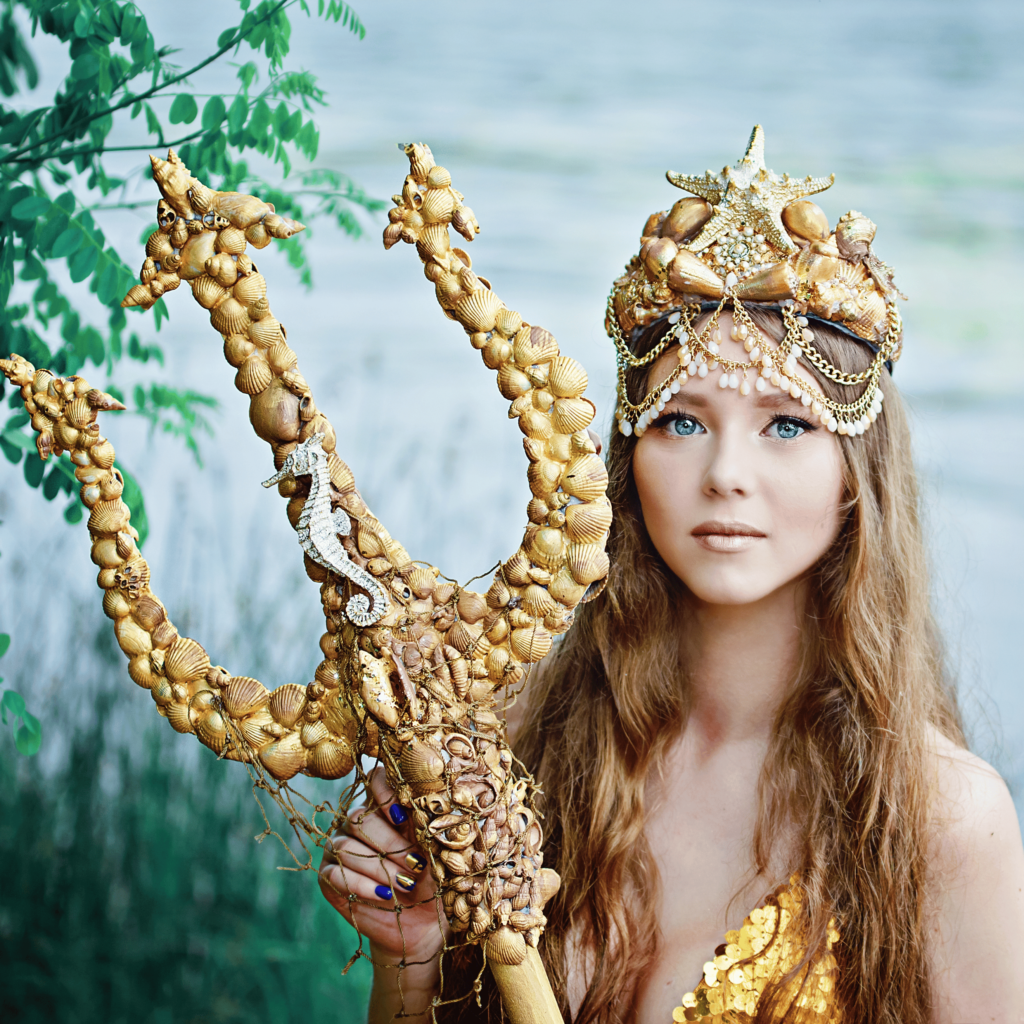 A woman dressed as a mermaid holds a trident adorned with seashells, embodying the benefits of natural serenity. She wears a shell and starfish crown and a sequined top, standing near water with a backdrop of greenery reminiscent of calming Reiki therapy.