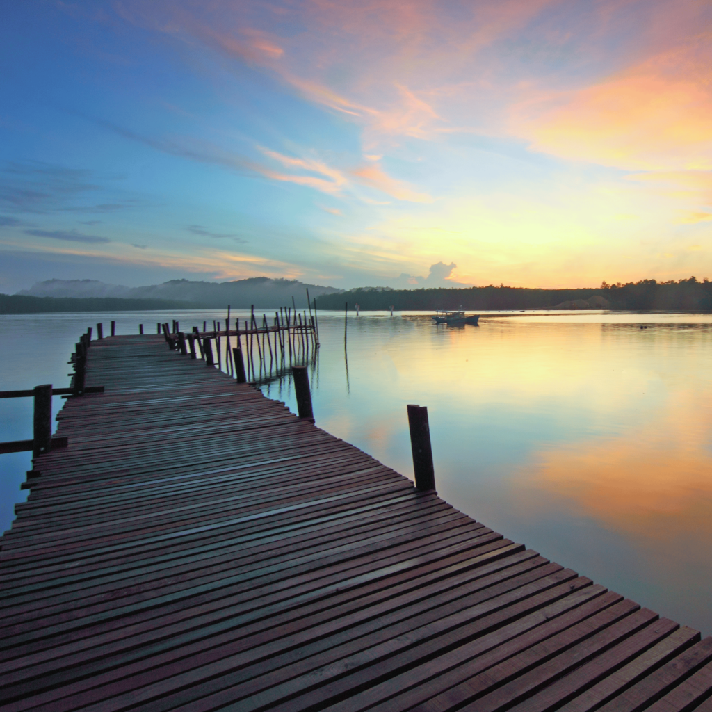A wooden pier extends over a calm lake at sunset, with vibrant orange, pink, and blue hues painting the sky and distant hills in the background. The serene scene feels like nature’s own Reiki Therapy, soothing to the soul as if crafted for chronic pain management.