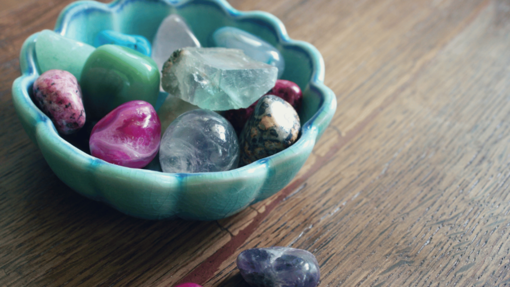 A blue ceramic bowl containing colorful polished stones—Reiki crystals for your healing journey—sits on a wooden surface, with an additional stone beside the bowl.